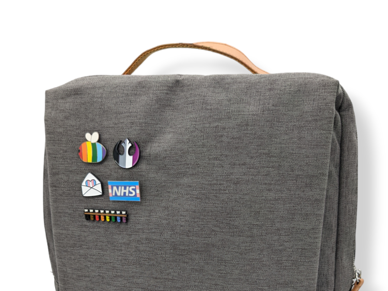 The top of a grey backpack that has five LGBTQ+ themed pin badges attached to it.